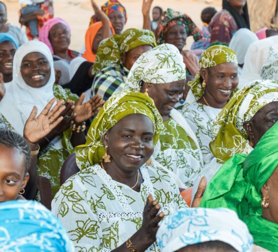 A large group of people gathered outdoors, seated closely together and wearing clothing with vibrant patterns and bright colors.