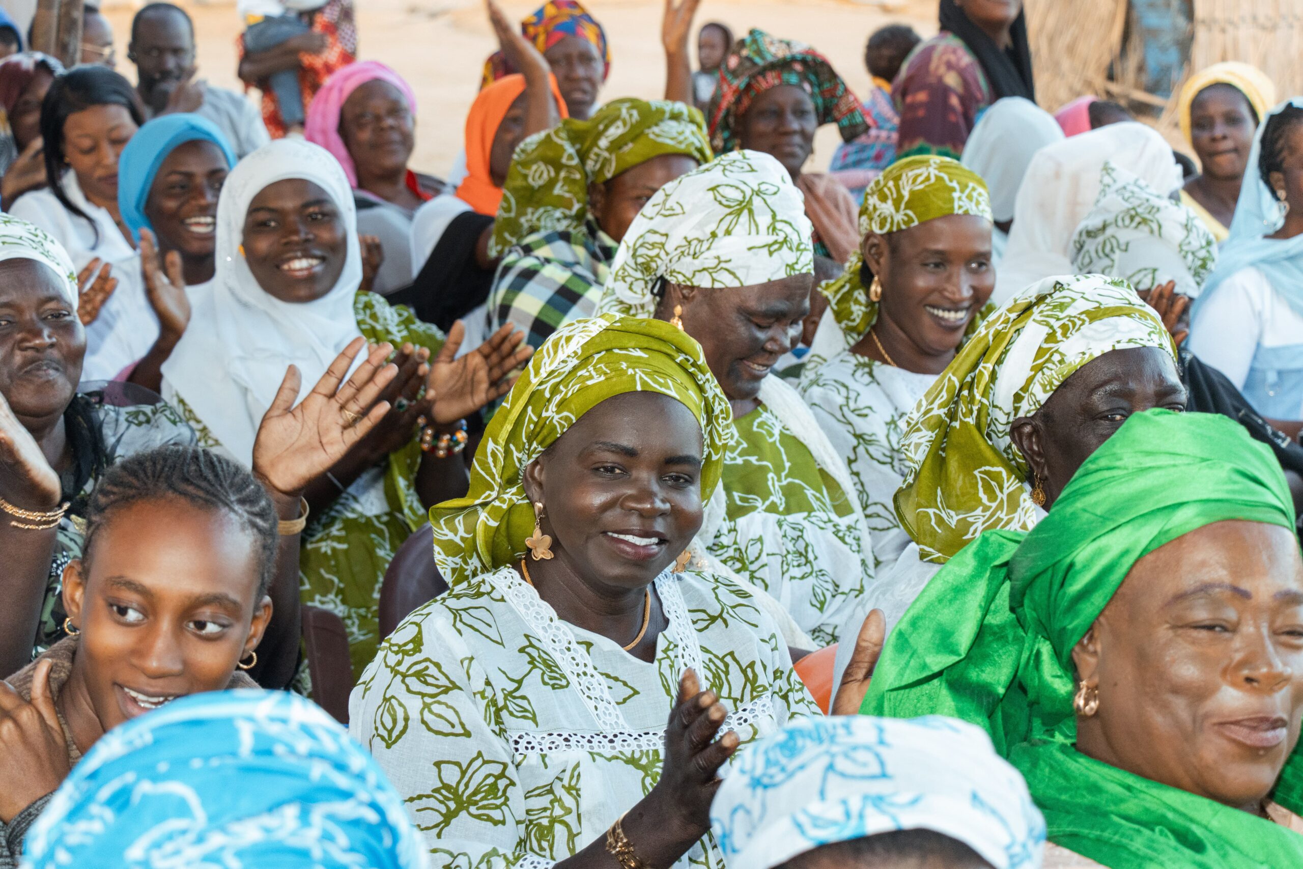A large group of people gathered outdoors, seated closely together and wearing clothing with vibrant patterns and bright colors.