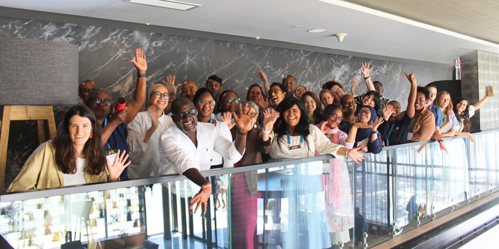 A large group of people standing together on an indoor balcony, raising their hands in a celebratory pose.