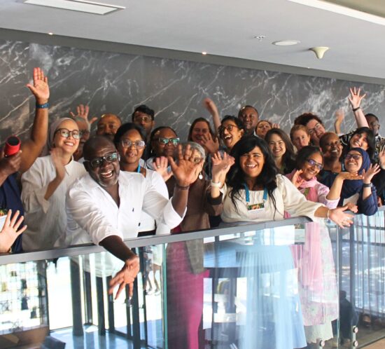 A large group of people standing together on an indoor balcony, raising their hands in a celebratory pose.